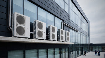 Row of large industrial air-conditioning chillers and condenser units on the rooftop of a British commercial building, with a hazy London office skyline in the background.
