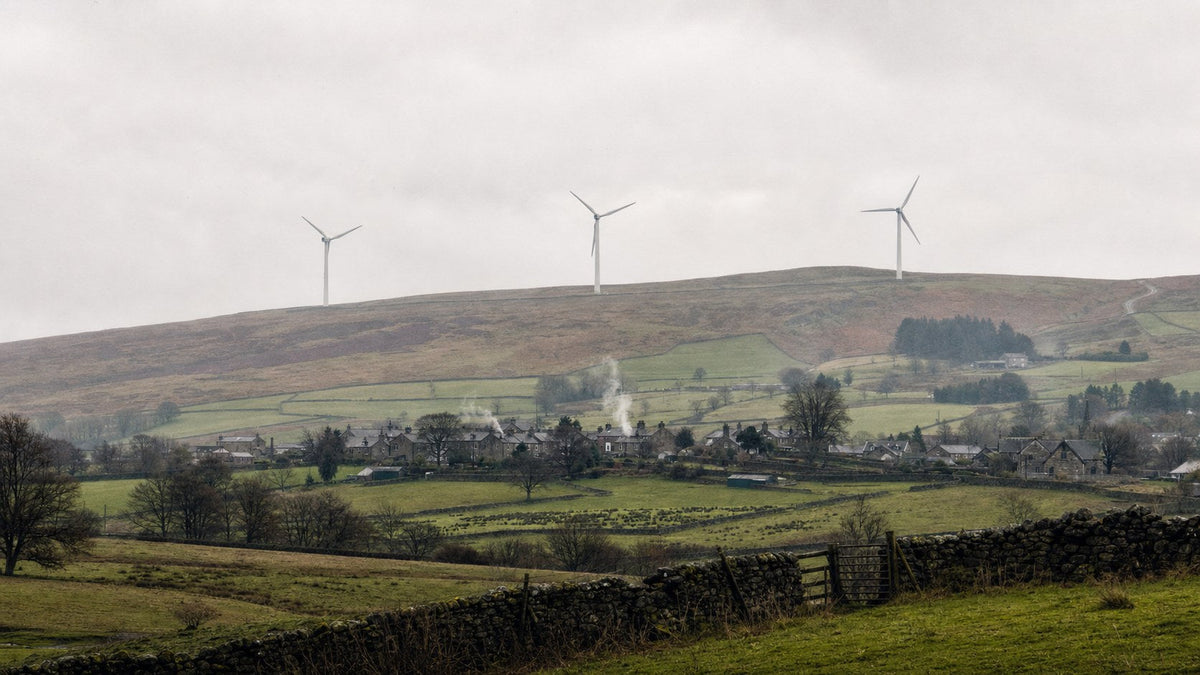 Three modern wind turbines on a hillside above a stone village in the British countryside on an overcast day, dry stone walls in the foreground